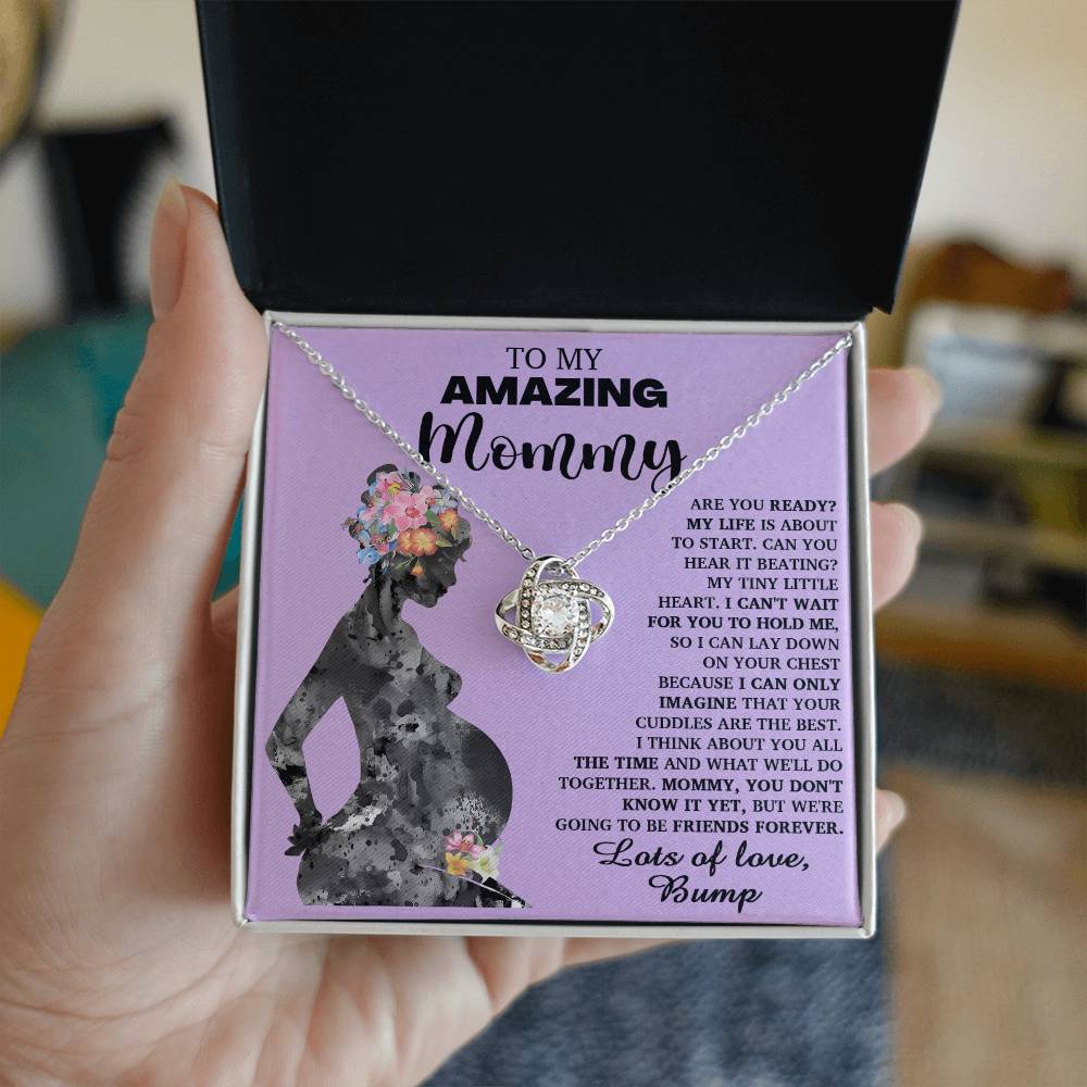A hand holds an open gift box containing the "To Mom To Be, Tiny Little Heart - Love Knot Necklace" adorned with premium cubic zirconia crystals. The inside of the box reads, "To my amazing mommy," with a heartfelt message. The illustration depicts a pregnant woman with a floral silhouette, symbolizing their unbreakable bond.