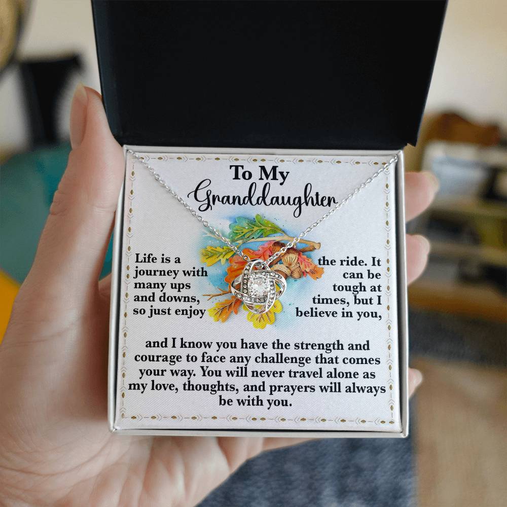 A person holds an open jewelry box, unveiling the Granddaughter-Never Travel Alone - Love Knot Necklace, beautifully embellished with shimmering Cubic Zirconia Crystals and accompanied by a heartfelt message for their granddaughter.