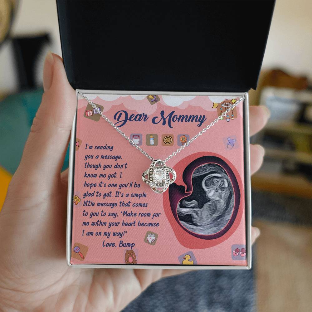 A hand holds a jewelry box containing the "To Mom To Be, On My Way - Love Knot Necklace," adorned with cubic zirconia crystals, placed on a note addressed to "Dear Mommy," featuring an image of an ultrasound and a heartfelt message from an unborn baby.