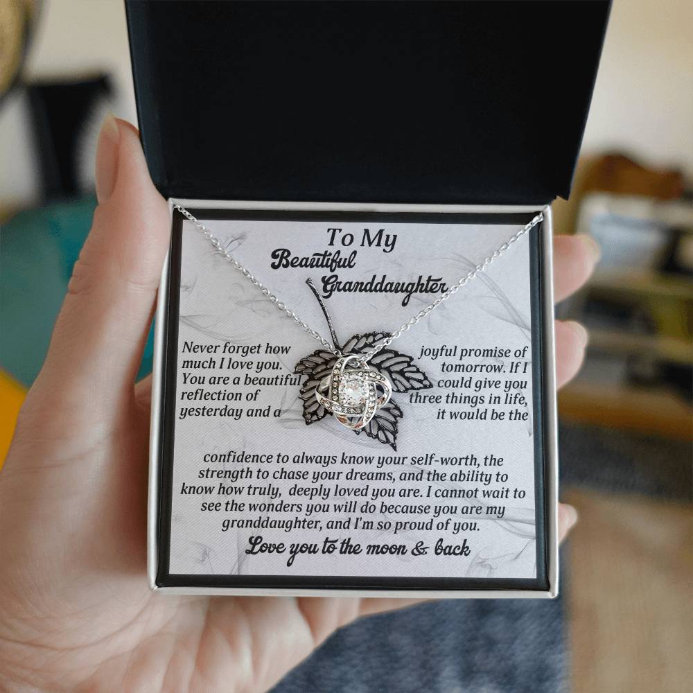 A person holds an open necklace box, showcasing the Granddaughter-Deeply Loved - Love Knot Necklace, embellished with cubic zirconia crystals. The pendant, finished in white gold, shares a heartfelt message meant for a granddaughter.