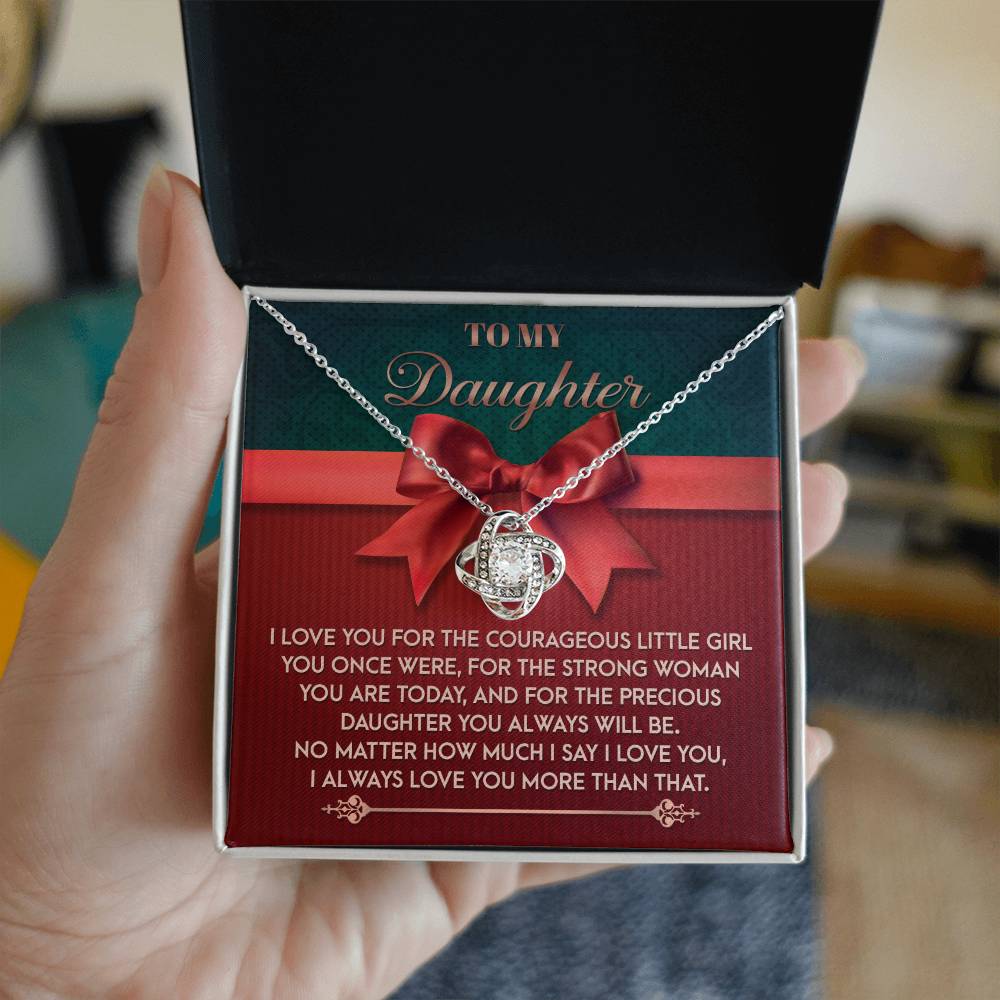 A hand holds a jewelry box, unveiling the Daughter-Always Will Be - Love Knot Necklace, beautifully embellished with cubic zirconia crystals. Inside, there's a card with a heartfelt message for a daughter.