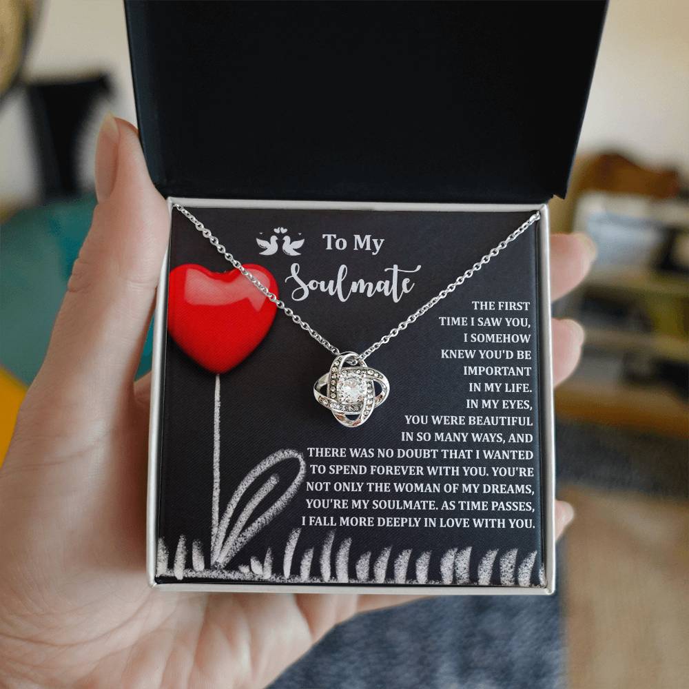 A hand holds a Soulmate-Forever With You - Love Knot Necklace gift box featuring a white gold finish with cubic zirconia crystals. Inside is a heartfelt message to a soulmate, adorned with a red heart and white floral illustration.