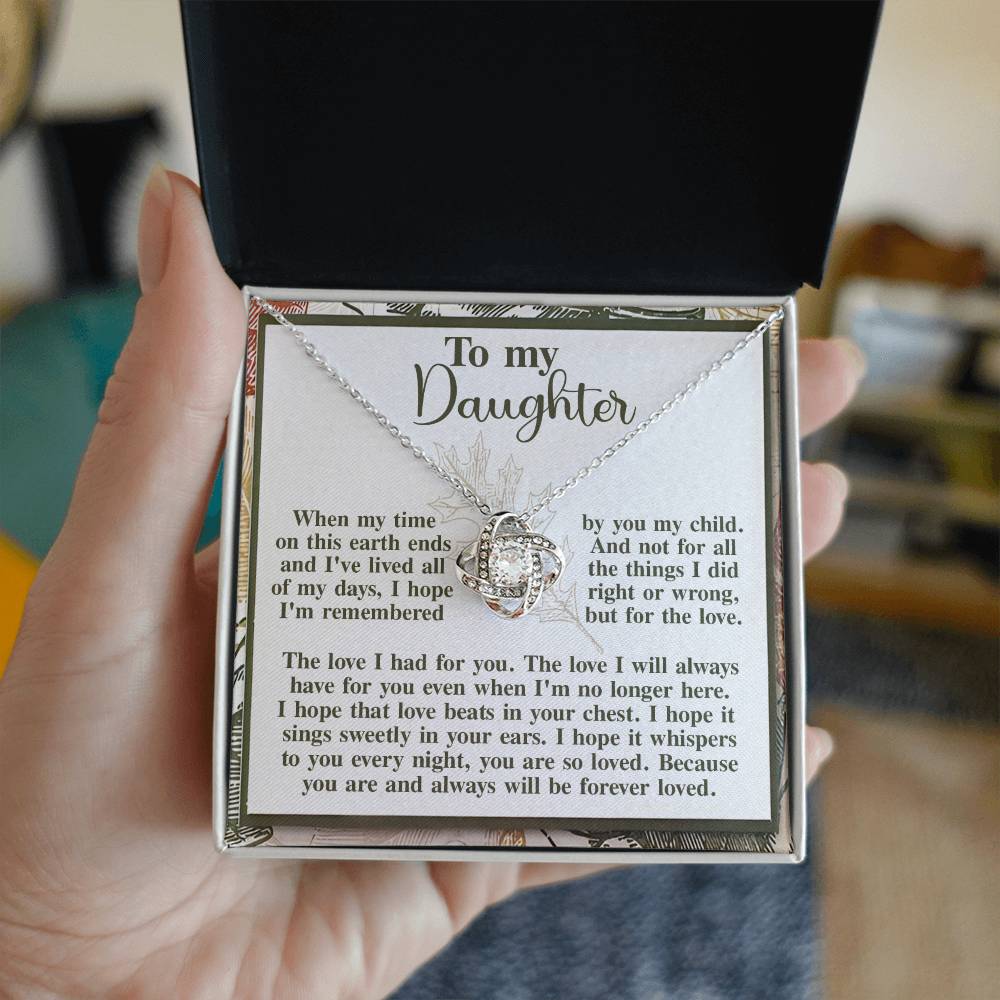 A hand holding an open box reveals a Daughter - In Your Ears Love Knot Necklace adorned with cubic zirconia crystals and a heartfelt note addressed "To my Daughter," with a message of everlasting love and remembrance. This personalized gift is truly special.