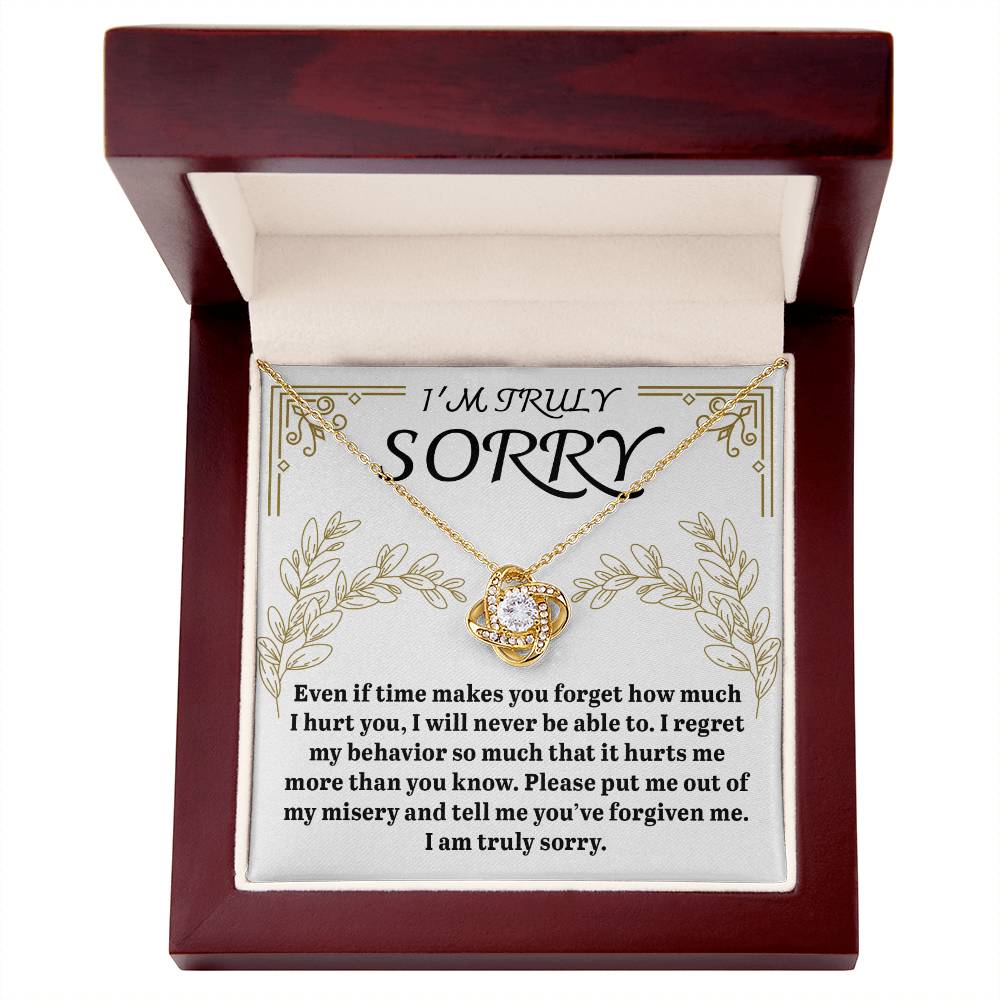 The "Sorry-It Hurts Me - Love Knot Necklace" features a circular pendant adorned with cubic zirconia crystals, elegantly displayed in an open wooden box. The box's lid presents an apology message that expresses deep regret and requests forgiveness.