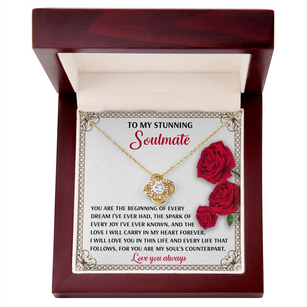 The Soulmate-Soul's Counterpart Love Knot Necklace, embellished with cubic zirconia, is elegantly displayed in a jewelry box. Its gold-finished circular pendant gleams under the lid adorned with roses and a romantic soulmate message.