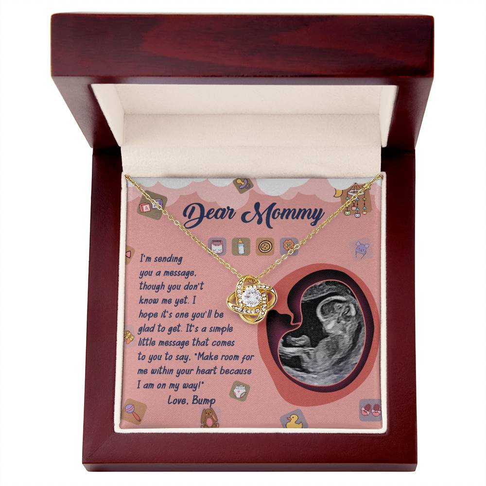 A personalized gift, the "To Mom To Be, On My Way - Love Knot Necklace," is showcased in an open wooden box. Inside the box is a card featuring a sonogram image and text addressed to "Dear Mommy," along with a heartfelt message from "Love, Bump," embellished with sparkling cubic zirconia crystals.