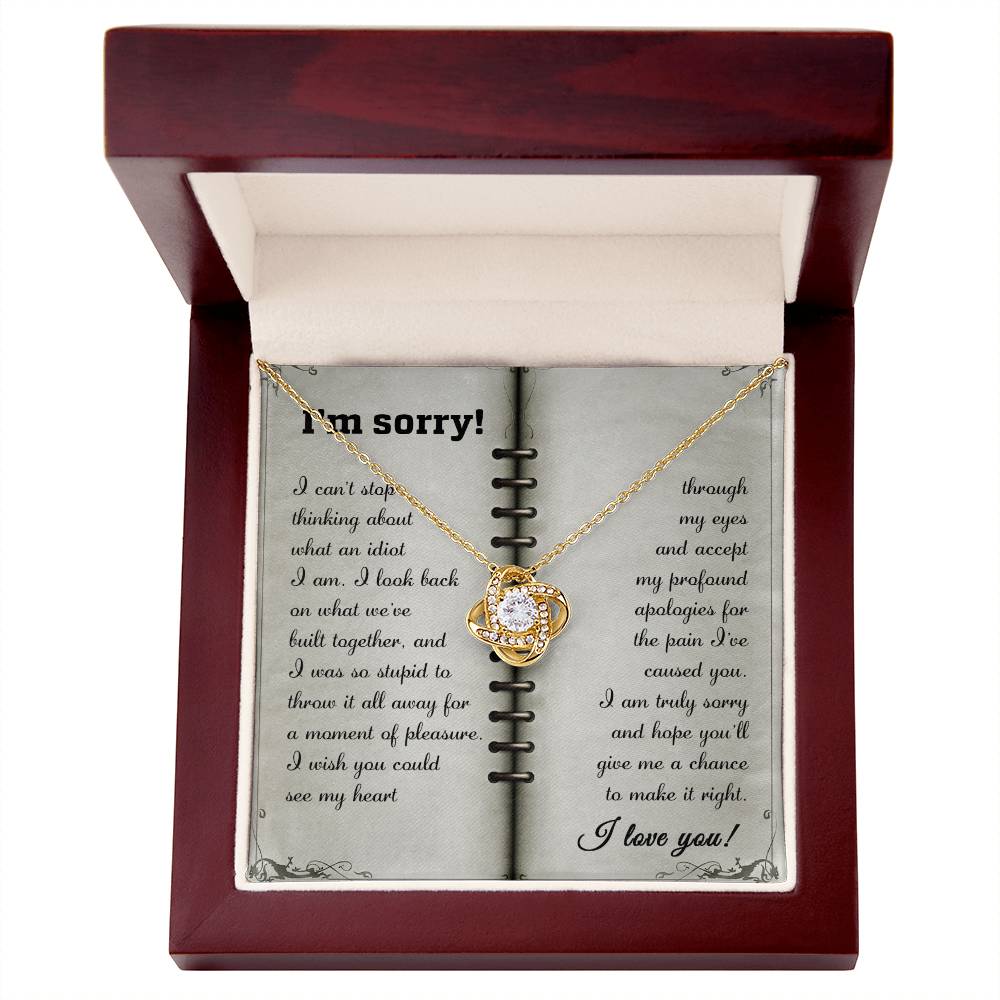 An open wooden box displays the "Sorry-What An Idiot - Love Knot Necklace" adorned with premium cubic zirconia crystals, placed over a card with an apology message ending with the words "I love you.