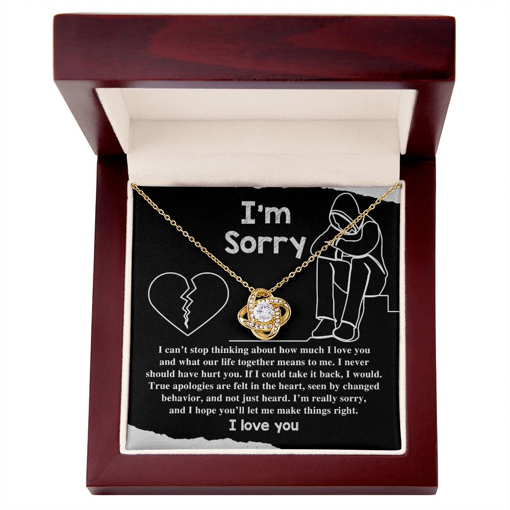 A gold Sorry-Not Just Heard Love Knot Necklace sits in an open wooden box, bearing a heartfelt message: "I'm sorry..." followed by a longer apology and expression of love. This personalized gift, enhanced with premium cubic zirconia crystals, conveys both elegance and deep emotion.