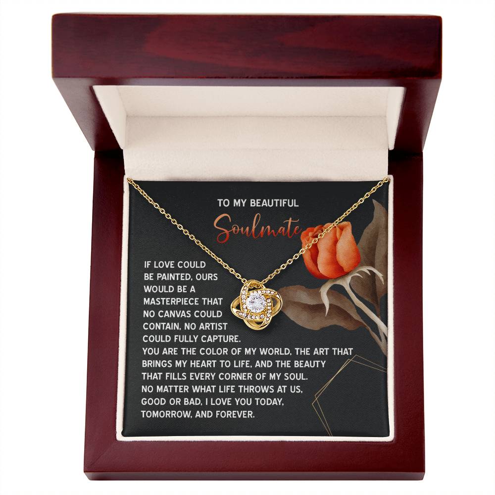 The Soulmate-A Masterpiece - Love Knot Necklace, featuring a central jewel and gold finish, is elegantly displayed in an open wooden box. Accompanying it is a card adorned with two roses and a romantic message that enhances its charm.