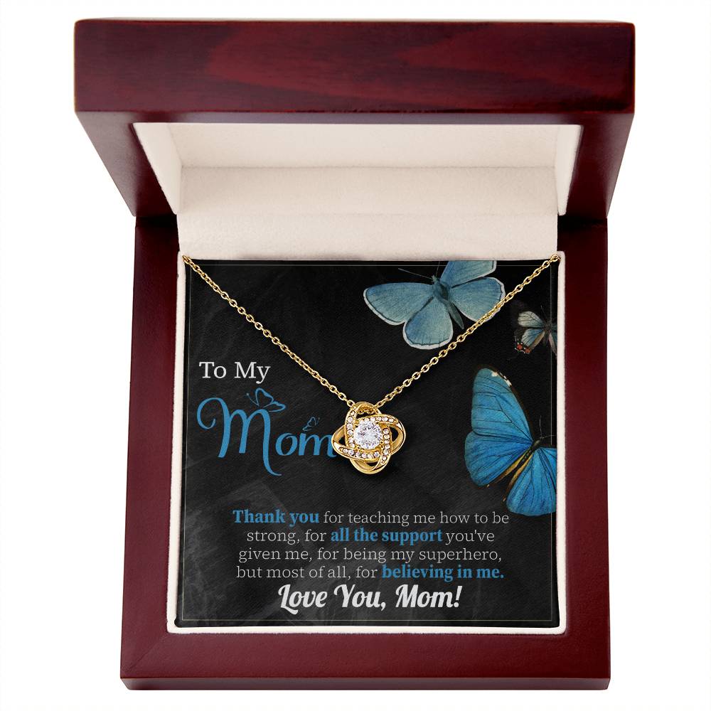 The "To Mom, Believing In Me - Love Knot Necklace" features a gold necklace with a cubic zirconia crystal pendant presented in a wooden gift box. The inside lid is adorned with blue butterfly images and words of appreciation, dedicated to 'Mom' for her support and love, making it the perfect personalized gift.