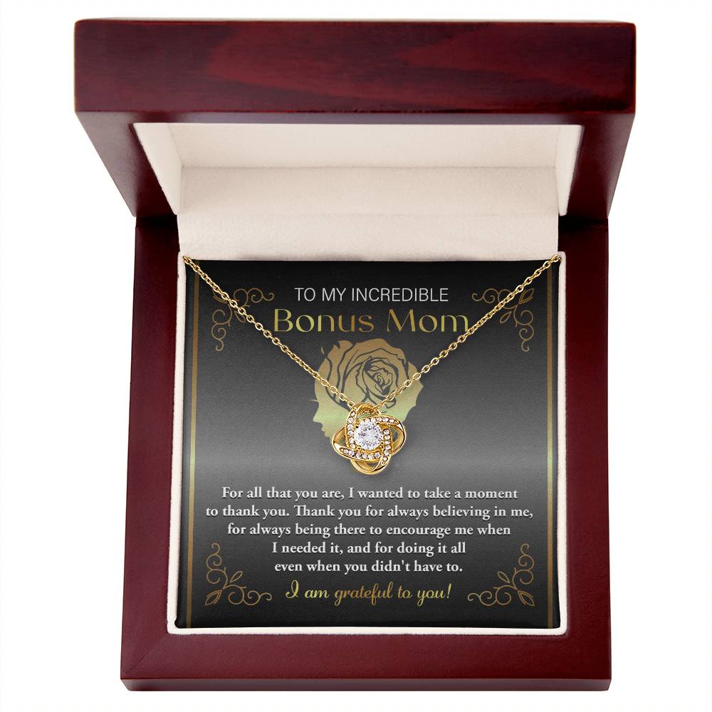 The "To Bonus Mom, To Encourage Me - Love Knot Necklace," featuring a pendant adorned with sparkling cubic zirconia crystals, is displayed in an open box. The inside of the box lid showcases a heartfelt message dedicated to a "Bonus Mom.