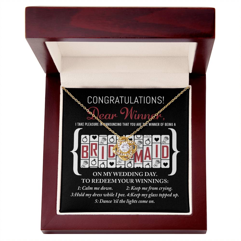 The "To Bridesmaid, The Winner Of - Love Knot Necklace" is displayed inside an open wooden box. The inside lid features a message with "BRIDESMAID" written prominently in the center, congratulating the recipient for being chosen and listing five tasks typically expected from a bridesmaid. The necklace sparkles with cubic zirconia crystals.