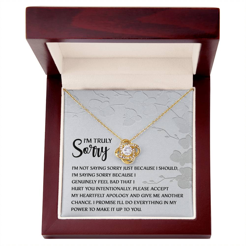 Introducing the Sorry-Feel Bad - Love Knot Necklace, featuring a pendant on a card with an apology message: "I'm truly sorry. I'm not saying sorry just because I should..." and continues with more heartfelt text. The necklace showcases a sparkling cubic zirconia crystal set in your choice of white gold or yellow gold finish, all against a light floral design background.