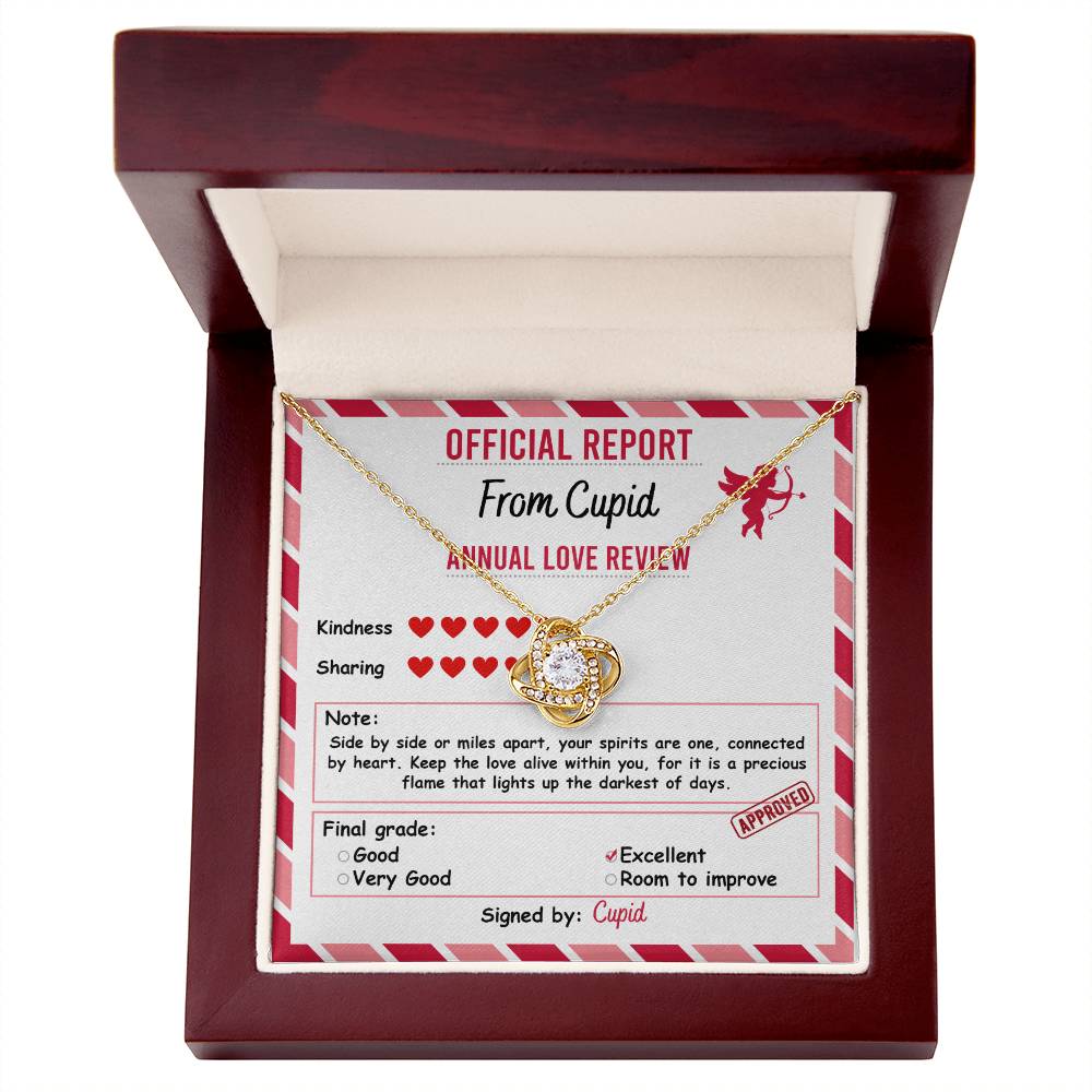 The Soulmate-Official Report's Love Knot Necklace features a heart pendant resting on a note in a wooden box. The "Annual Love Review" note has checkboxes marked "Excellent" and is adorned with cubic zirconia crystals, signed by "Cupid.