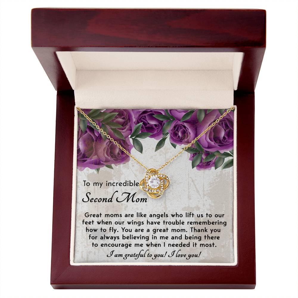 The "To Bonus Mom, Grateful For You - Love Knot Necklace," featuring a gold pendant and a central gemstone, is displayed in an open wooden box. Adorned with sparkling cubic zirconia crystals, it bears a heartfelt message for a "second mom," all set against a backdrop of purple flowers. This personalized gift is truly one-of-a-kind.