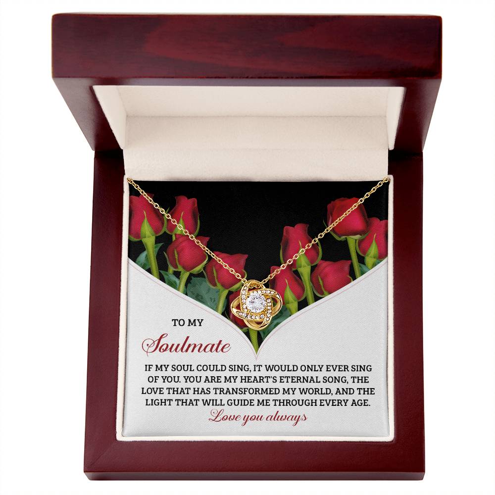 A Soulmate-Eternal Song Love Knot Necklace in white or yellow gold rests elegantly over roses in an open wooden jewelry box, harmonizing with the romantic soulmate message etched inside the lid.