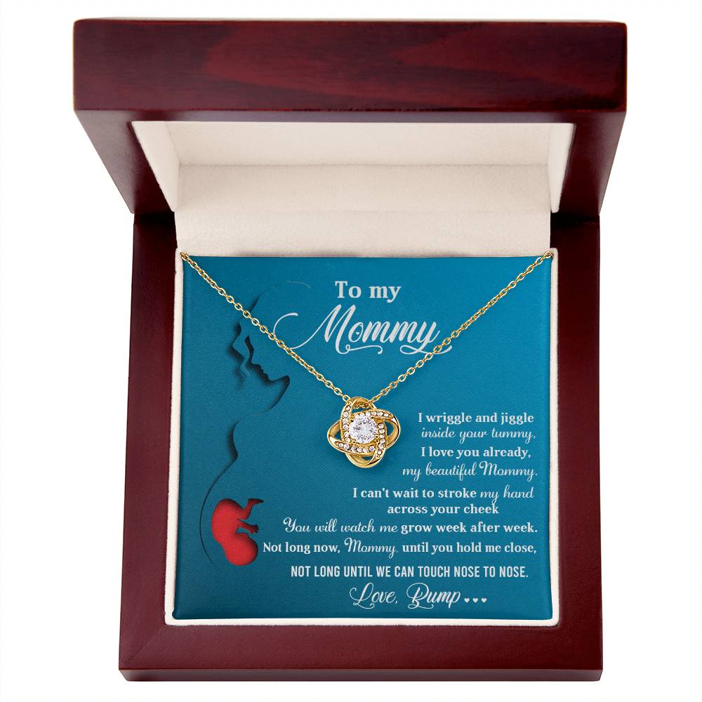 The “To Mom To Be, Nose To Nose - Love Knot Necklace,” crafted from 14k white gold and adorned with cubic zirconia crystals, is displayed in a wooden box. The box lid contains a heartfelt message to "Mommy" from "Bump," expressing love and anticipation of the baby's arrival.
