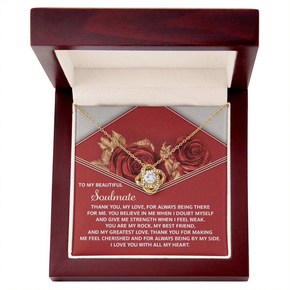 The Soulmate-All My Heart Love Knot Necklace features a gold heart-shaped design with a gemstone and cubic zirconia crystals, presented in a red box. The lid displays an inspiring message about love and support, framed by elegant rose patterns.