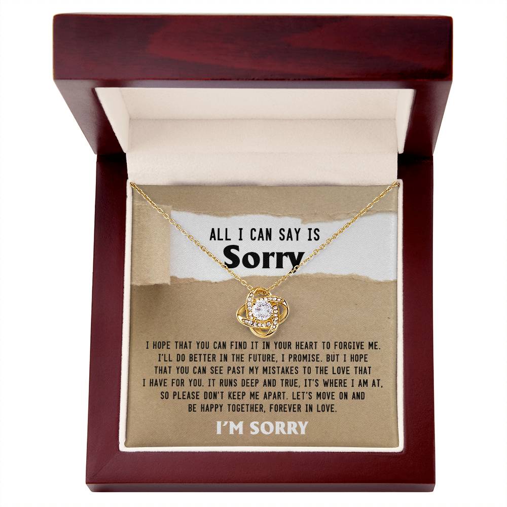 A **Sorry-Keep Me Apart - Love Knot Necklace** with a circular pendant adorned with cubic zirconia crystals is displayed in an open wooden box. Inside the box lid, a printed apology message is visible that emphasizes hope for forgiveness and expresses regret.