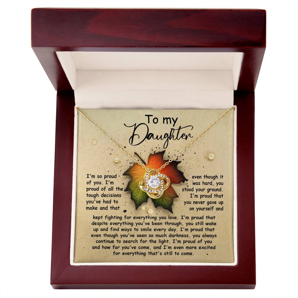 The "Daughter-Stood Your Ground - Love Knot Necklace" with a gold finish and pendant is elegantly showcased in a box. The box features a heartfelt message addressed "To my Daughter," expressing pride, admiration, and encouragement. Sparkling cubic zirconia crystals adorn the necklace, adding an extra touch of elegance.
