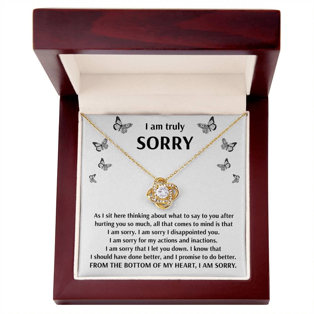 The Sorry-Done Better - Love Knot Necklace, adorned with cubic zirconia crystals, rests inside an open wooden box. The lid of the box features an apology message printed on white fabric, surrounded by small butterfly images—making it a truly personalized gift.