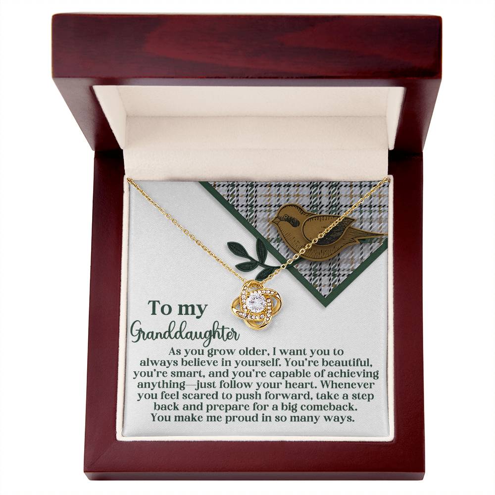 Open wooden box featuring a heartfelt message card for a granddaughter and the Granddaughter-A Big Comeback - Love Knot Necklace adorned with a gold-tone floral pendant intertwined with sparkling Cubic Zirconia.
