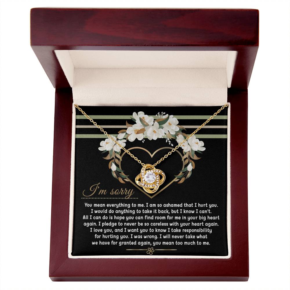 Introducing the Sorry-Big Heart - Love Knot Necklace: a heart-shaped pendant nestled in an elegant box, complete with an apology message expressing love and regret for causing hurt. The backdrop showcases a beautiful floral heart design, while the pendant sparkles with cubic zirconia crystals. Personalize your necklace by choosing between a white gold or yellow gold finish.