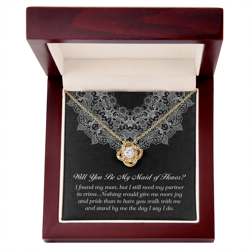 A gold Love Knot Necklace with a flower-shaped pendant containing cubic zirconia crystals is displayed in a dark red wooden box. The box has an inscription on black velvet, asking, "Will You Be My Maid of Honor?" with a heartfelt message about partnership and joy.