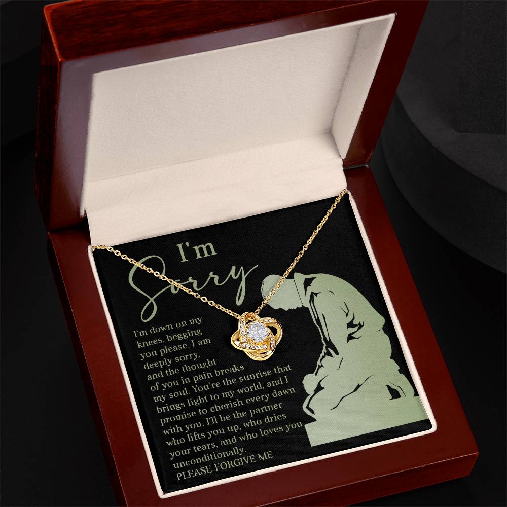 The "Sorry-On My Knees - Love Knot Necklace" is a gold necklace with a heart-shaped pendant, presented in a wooden box. Enhanced with cubic zirconia crystals, the box interior features an apology message and an image of a kneeling figure in green.
