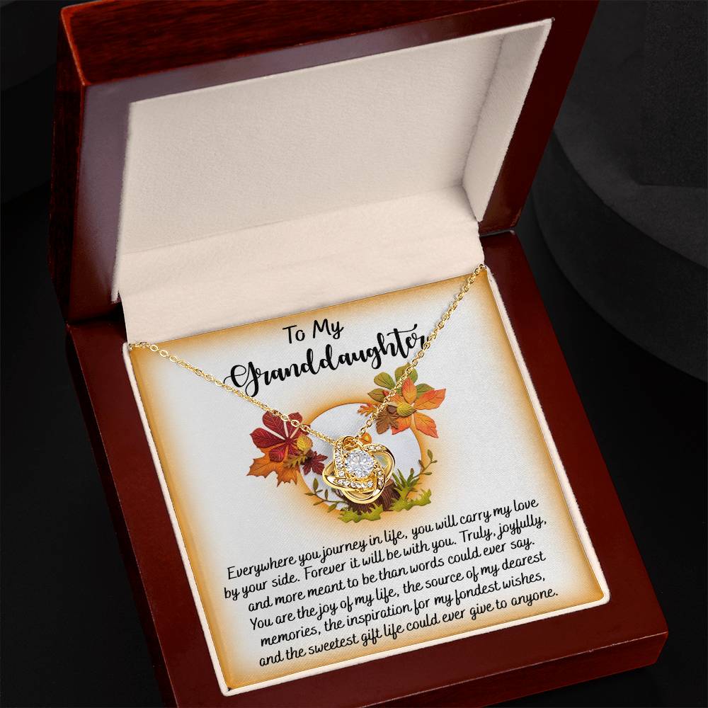 The Granddaughter-By Your Side - Love Knot Necklace, featuring sparkling cubic zirconia crystals and adorned with a leaf and heart pendant, is elegantly displayed in a wooden box. Available in both white gold or yellow gold finish, the inside lid holds a heartfelt message dedicated to a beloved granddaughter.