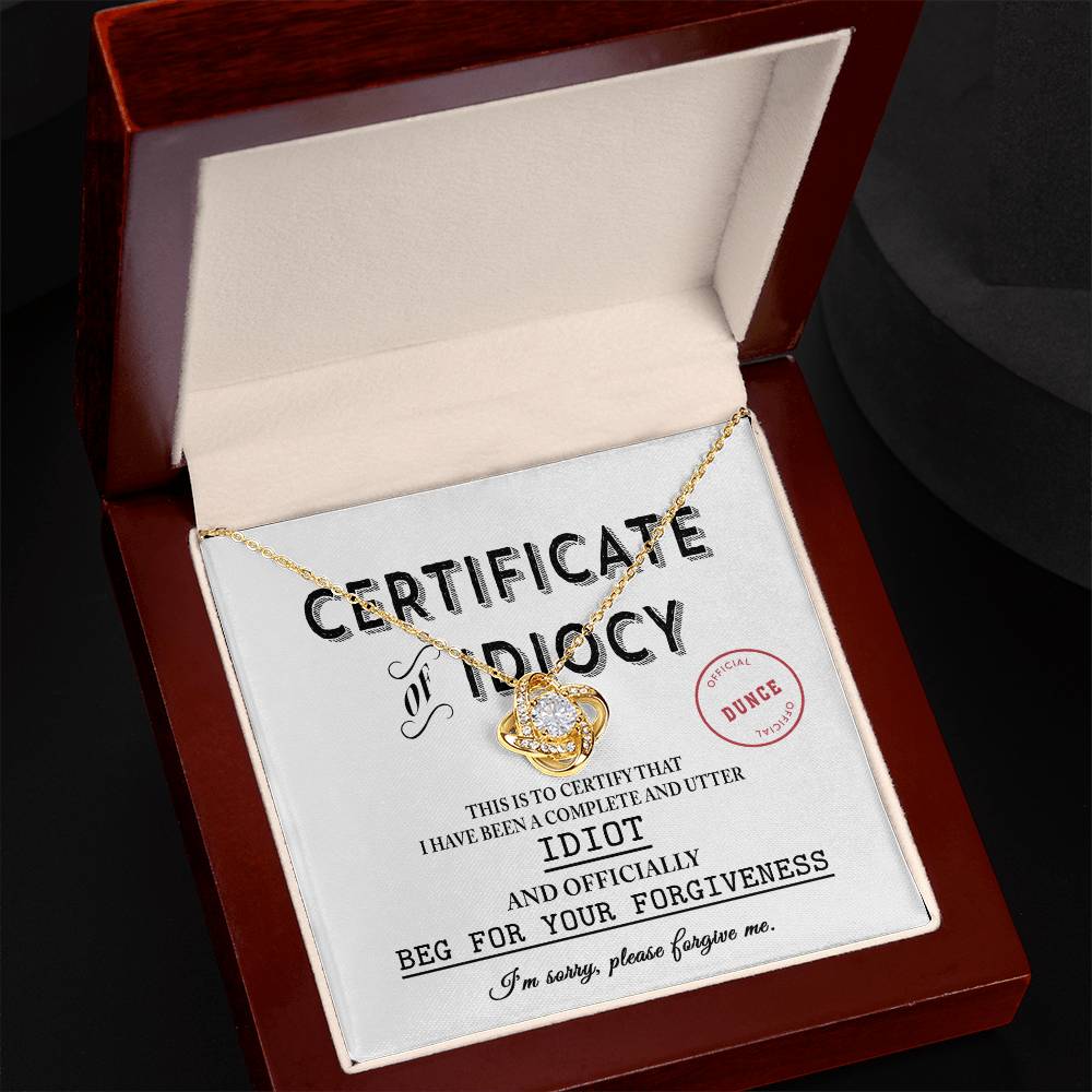 A jewelry box containing a Sorry-Certificate Of Idiocy - Love Knot Necklace adorned with cubic zirconia crystals rests atop a certificate titled "Certificate of Idiocy," which apologizes for being an idiot and requests forgiveness.
