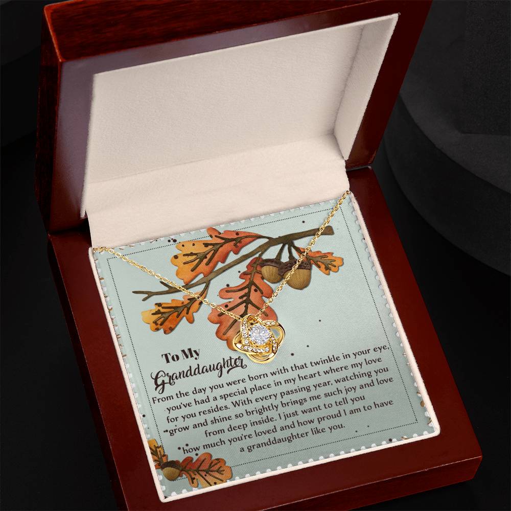 The "Granddaughter-From Deep Inside - Love Knot Necklace" features a radiant cubic zirconia that shines elegantly in a wooden box. The inside lid carries a heartfelt message to a granddaughter, beautifully framed against the backdrop of autumn leaves, creating an enchanting scene.