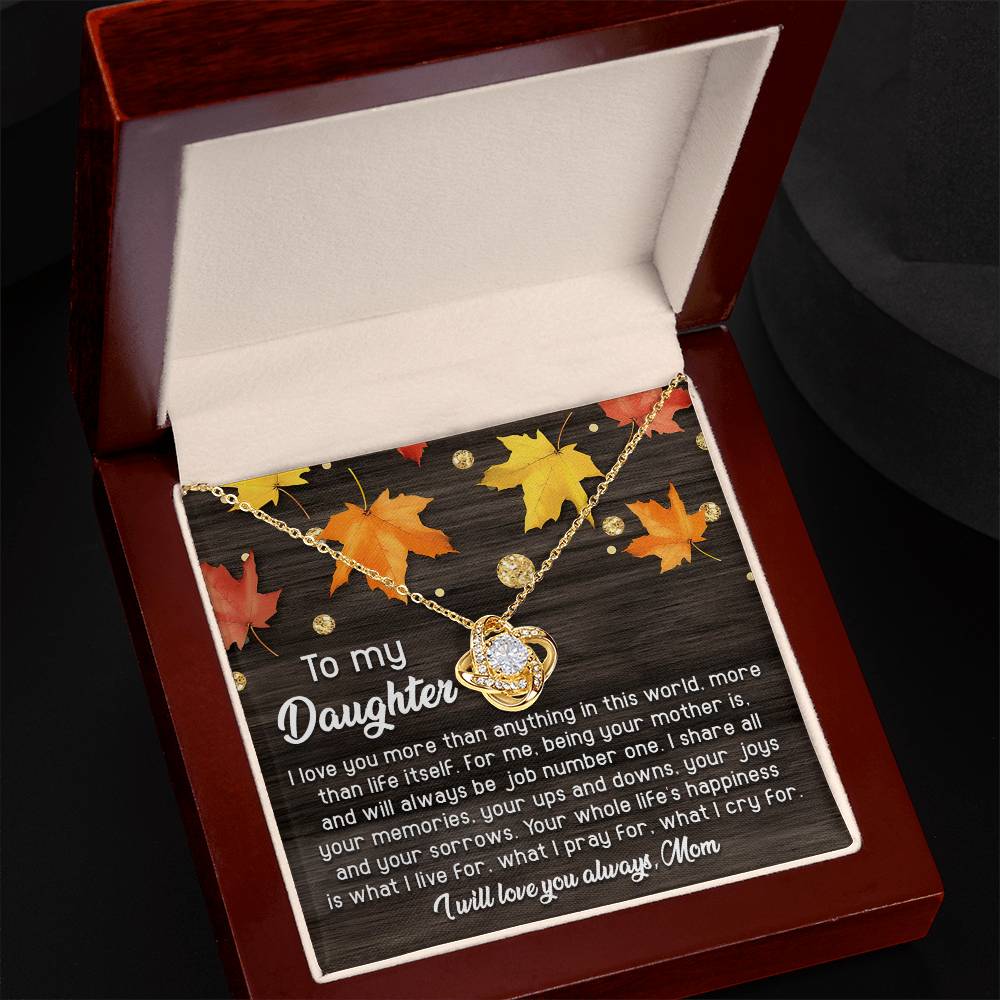 An open jewelry box showcases the Daughter-Job Number One - Love Knot Necklace, adorned with premium cubic zirconia crystals, on a dark, autumn-themed background. The inscribed message begins with "To my Daughter" and ends with "love you always, Mom." This personalized gift is truly heartfelt.