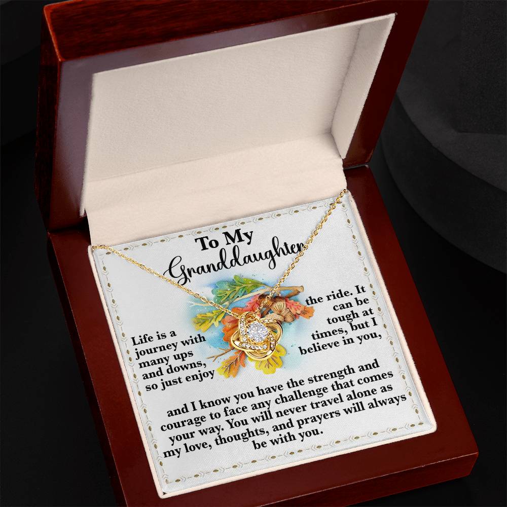 The Granddaughter-Never Travel Alone - Love Knot Necklace comes in a jewelry box with a heart pendant. The inside lid reveals a heartfelt message for a granddaughter, beautifully surrounded by flowers and inspiring words about life's journey. This elegant piece is adorned with cubic zirconia crystals that enhance its gold finish.