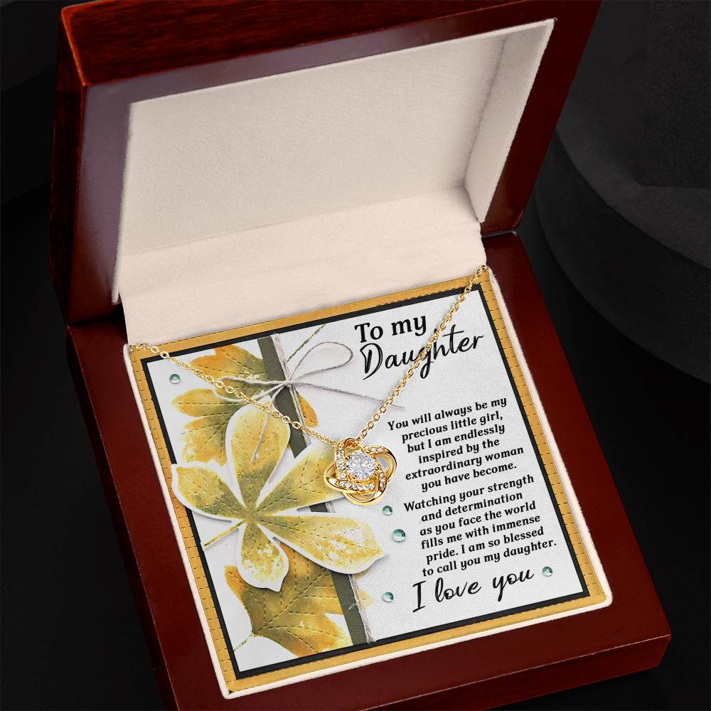 An open jewelry box reveals the stunning Daughter-Blessed To Call - Love Knot Necklace, featuring a heart pendant with an inspirational message dedicated to a daughter. The heartfelt message is elegantly surrounded by a yellow and white floral design, beautifully enhanced by the necklace's exquisite gold finish.