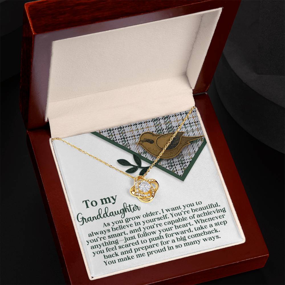 The Granddaughter-A Big Comeback - Love Knot Necklace, featuring interlocking hearts and a heartfelt message for a granddaughter, sparkles in its open jewelry box with elegant Cubic Zirconia Crystals and a radiant Gold Finish.