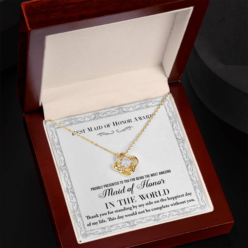 A Love Knot Necklace, elegantly crafted in gold and featuring a heart-shaped pendant adorned with shimmering cubic zirconia crystals, rests in an open jewelry box. Inside the box, a note reads: "To Maid of Honor, Of My Life - Best Maid of Honor Award. Proudly presented to you for being the most amazing Maid of Honor in the world. Thank you for standing by my side on the happiest day of my life.