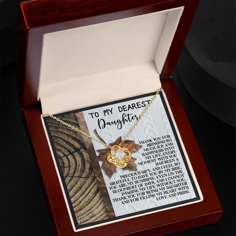 The Daughter-Love And Pride - Love Knot Necklace, featuring a gold pendant adorned with sparkling cubic zirconia crystals, is beautifully presented in an open jewelry box. It comes with a heartfelt message for a daughter, expressing love and pride. This classic gift embodies timeless elegance and deep affection.