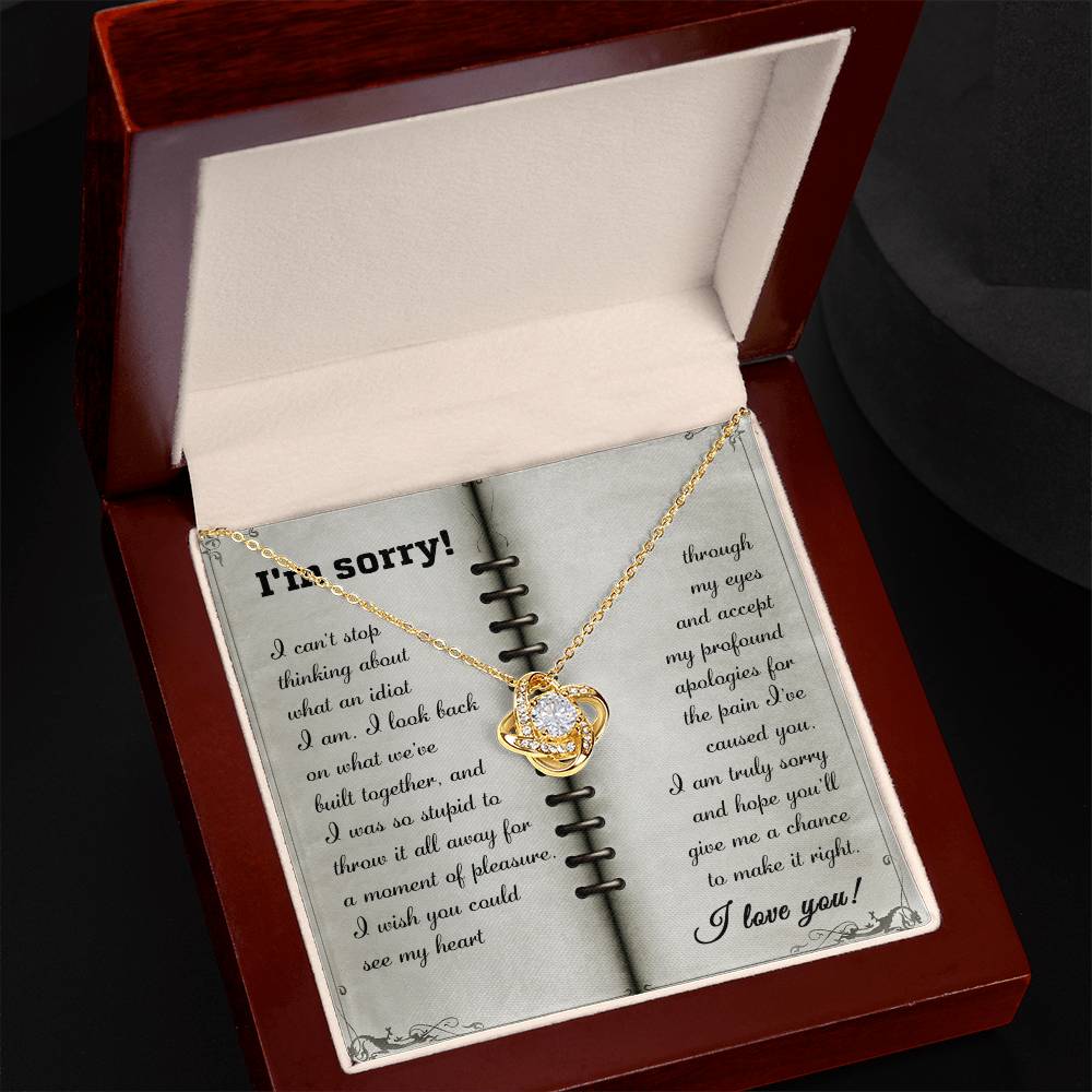 A gold heart-shaped pendant necklace with a crystal center, known as the "Sorry-What An Idiot - Love Knot Necklace," is displayed in an open jewelry box, accompanied by an apology message on an open book background—a perfect personalized gift.