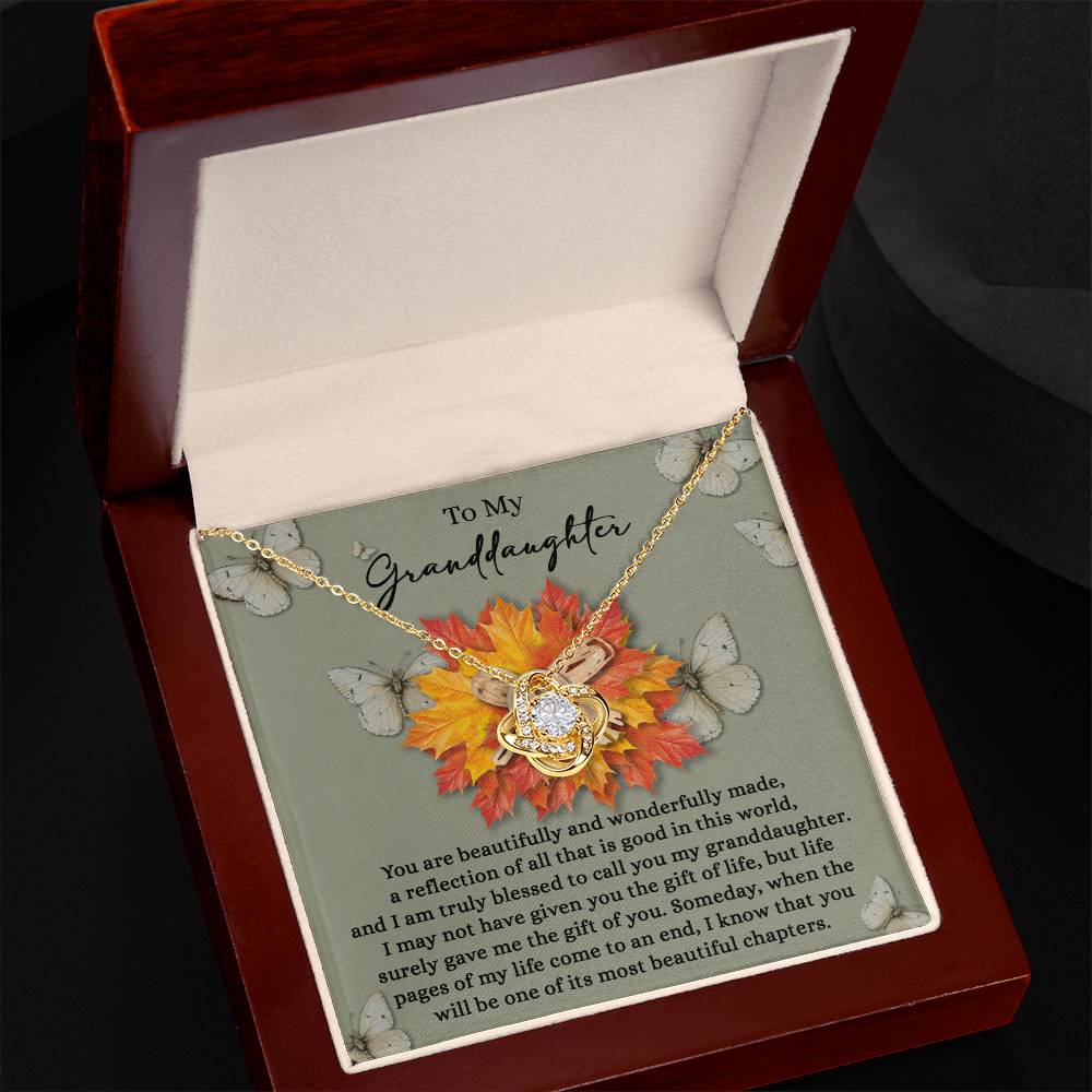 A Granddaughter-Gift Of You - Love Knot Necklace in a gold finish, featuring a circular pendant, is beautifully presented in a jewelry box with a heartfelt message for your granddaughter. The background is adorned with butterflies and autumn leaves, which glisten gently like cubic zirconia crystals in the light.