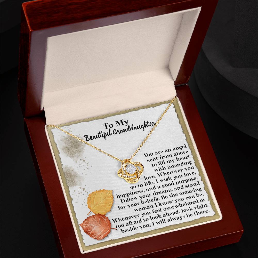 Presenting the Granddaughter-An Angel - Love Knot Necklace, nestled in an elegant jewelry box with a white gold finish. The necklace sparkles with cubic zirconia crystals, while inside the lid, a leafy design enhances the heartfelt message for a granddaughter, creating a treasured keepsake.