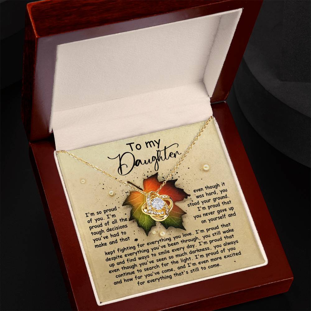 A Love Knot Necklace named "Daughter-Stood Your Ground," featuring a heart and infinity pendant adorned with cubic zirconia crystals, presented in a box with a sentimental message that reads "To my Daughter" against a colorful leaf design background.