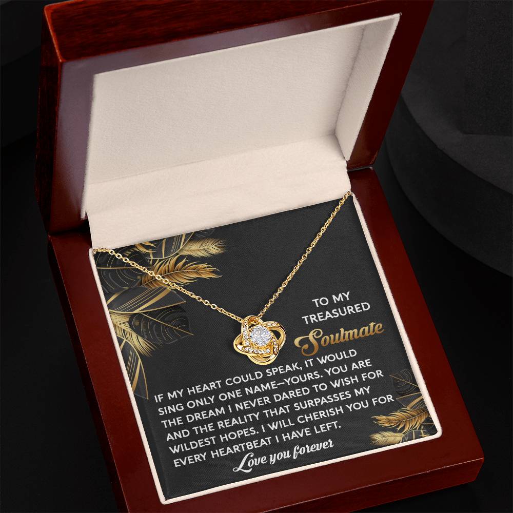 The Soulmate-Wildest Hopes Love Knot Necklace, featuring sparkling cubic zirconia crystals, lies in an open jewelry box designed for a cherished soulmate against a black and gold leaf backdrop.
