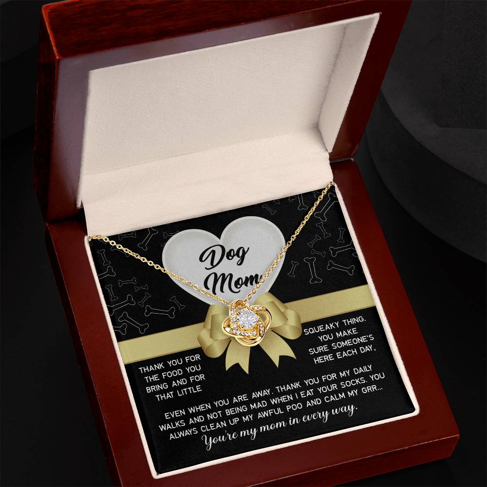The "Dog Mom-In Every Way" Love Knot Necklace features a gold intertwined heart pendant with cubic zirconia crystals. It comes in a wood and white velvet box, with the lid displaying "Dog Mom" alongside bone and heart graphics, expressing gratitude and love for a dog's caregiver.