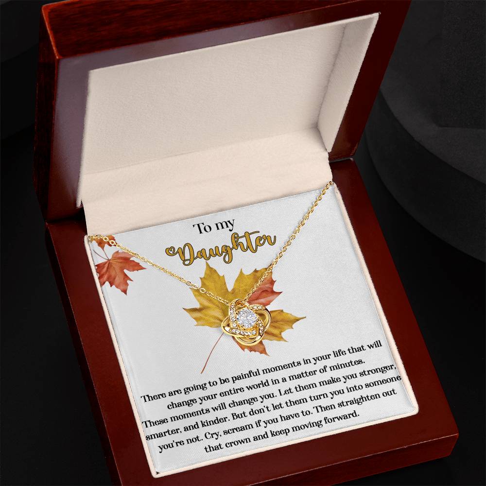 The "Daughter-Make You Stronger - Love Knot Necklace," a gold necklace with a cubic zirconia-adorned pendant, is displayed in an open wooden box. The box’s interior features an inscription starting with "To my Daughter" and includes decorative leaves.