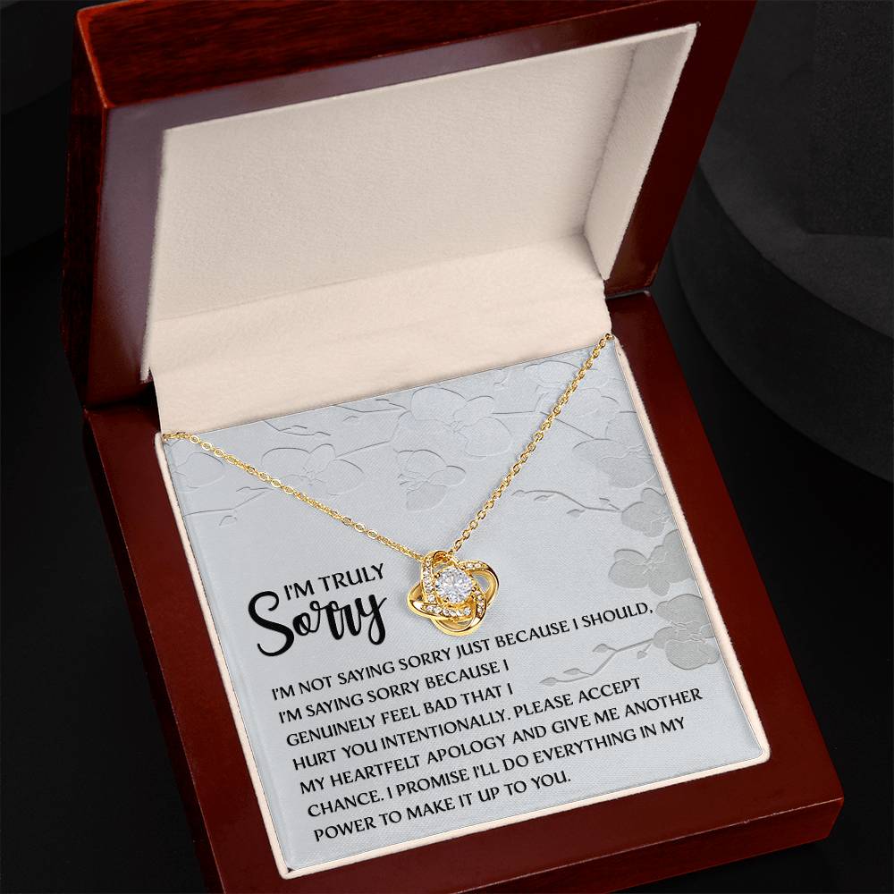 Introducing the Sorry-Feel Bad - Love Knot Necklace, featuring a pendant on a card with an apology message: "I'm truly sorry. I'm not saying sorry just because I should..." and continues with more heartfelt text. The necklace showcases a sparkling cubic zirconia crystal set in your choice of white gold or yellow gold finish, all against a light floral design background.