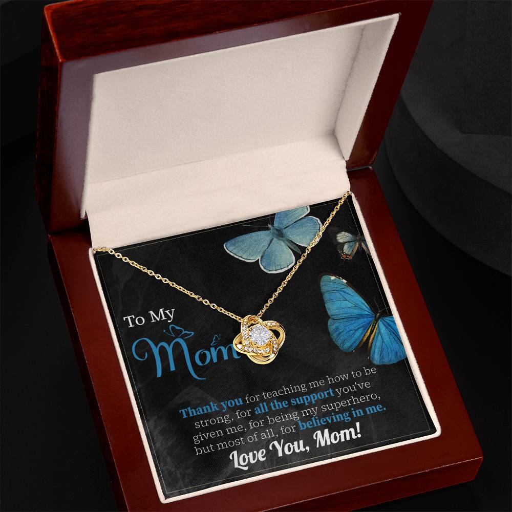 The "To Mom, Believing In Me - Love Knot Necklace" with a cubic pendant sits in an open wooden box on a note that reads: "To my Mom, Thank you for teaching me how to be strong... Love You, Mom!" adorned with blue butterfly illustrations. A truly personalized gift featuring cubic zirconia crystals.