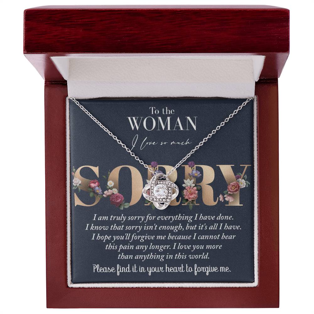 The Sorry-Bear This Pain - Love Knot Necklace, featuring a heart-shaped pendant adorned with cubic zirconia crystals and crafted in 14k white gold, is displayed in a red jewelry box. The interior of the lid has a heartfelt apology message for forgiveness written in decorative floral text.