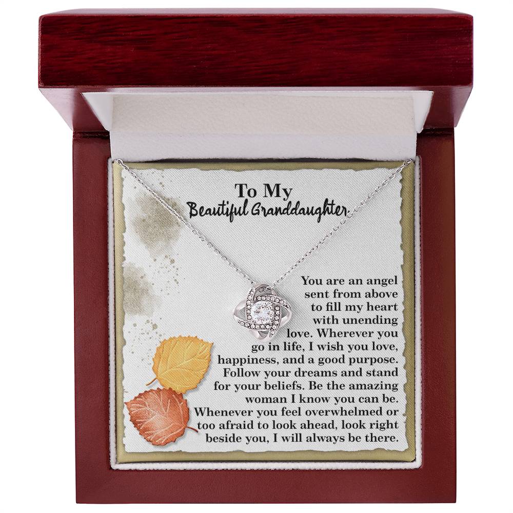 The Granddaughter-An Angel - Love Knot Necklace with cubic zirconia crystals is elegantly displayed in an open wooden box, adorned with a heartfelt message for a granddaughter and surrounded by decorative leaves.