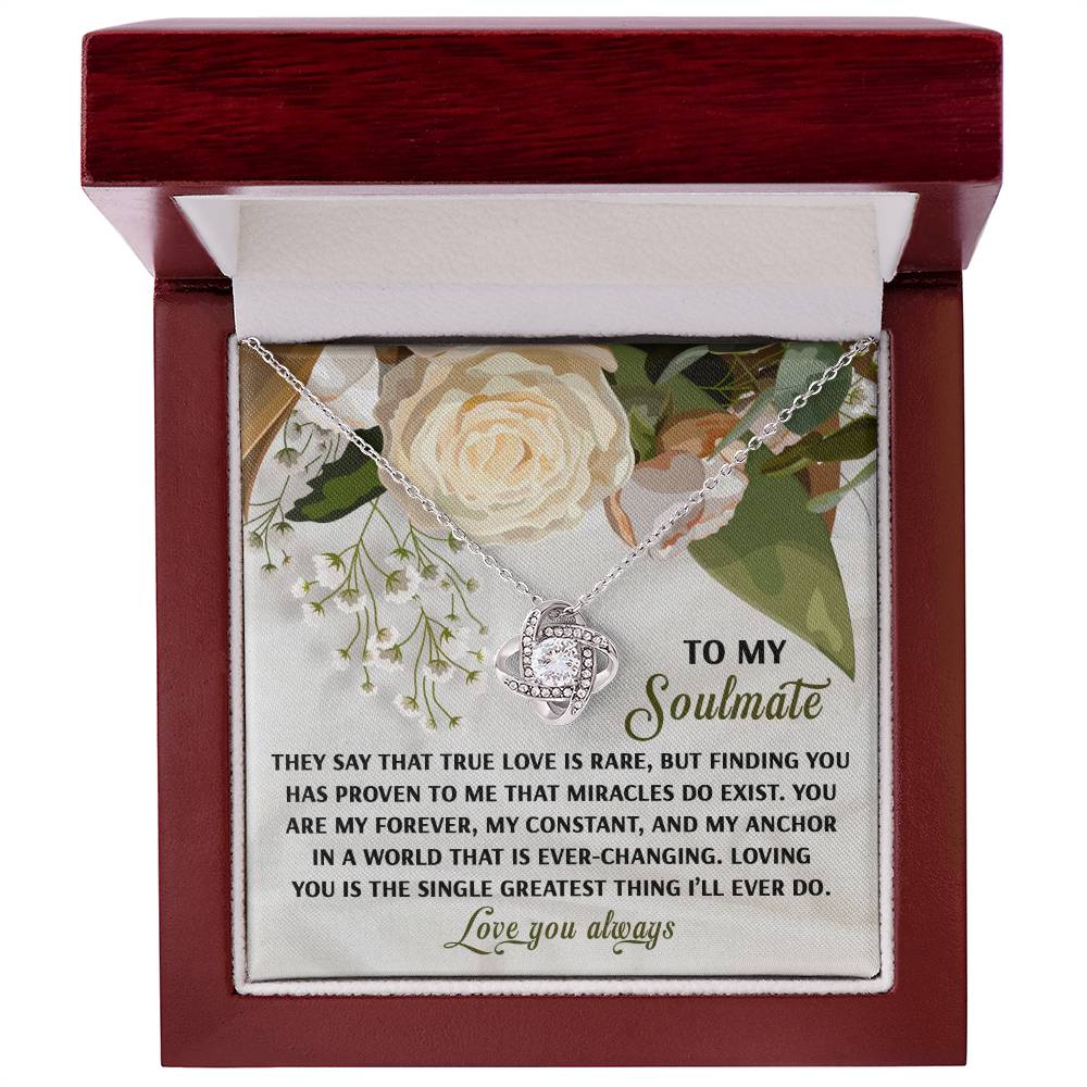 The Soulmate-Greatest Thing Love Knot Necklace features intertwined rings with cubic zirconia crystals, nestled in a box with a floral backdrop and a romantic soulmate message, available in white or yellow gold finish.